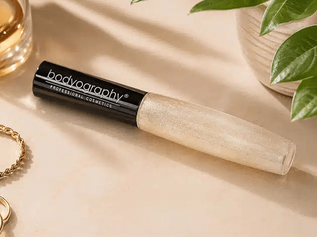 Bodyography professional cosmetics concealer tube with beige applicator on a light countertop, nearby green plant leaves.