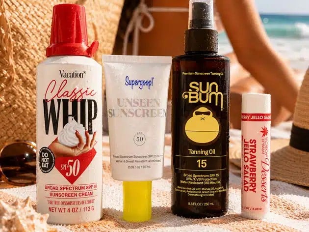 Assorted sunscreen bottles lined up on a sandy beach with the ocean in the background, including a red-capped spray, a white squeeze tube labeled 'Unseen Sunscreen', a dark bottle 'Sun Bum' spray, and a small SPF lip balm in the foreground—ready for sun protection on a sunny day.