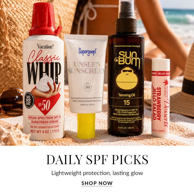 Assorted sunscreen bottles on a sandy beach, including SPF 50 cream, SPF 50 unseen sunscreen, SPF 15 tanning oil, and a lip balm; overlay reads Daily SPF Picks (advertisement).