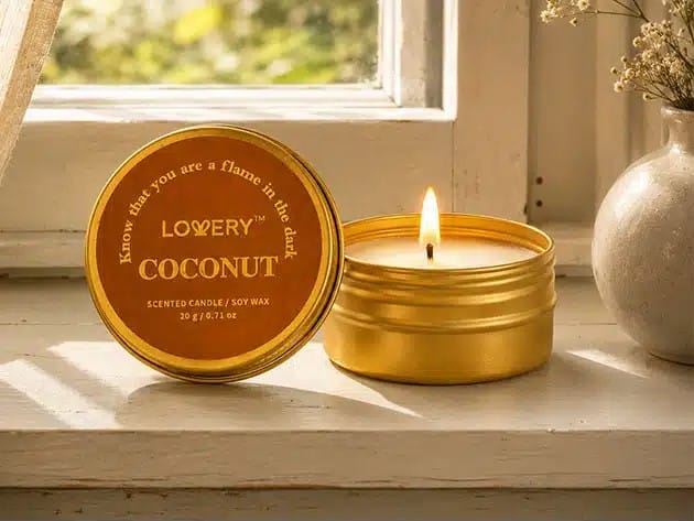 Gold tin candle labeled coconut with a lit wick on a sunlit windowsill, lid beside it.